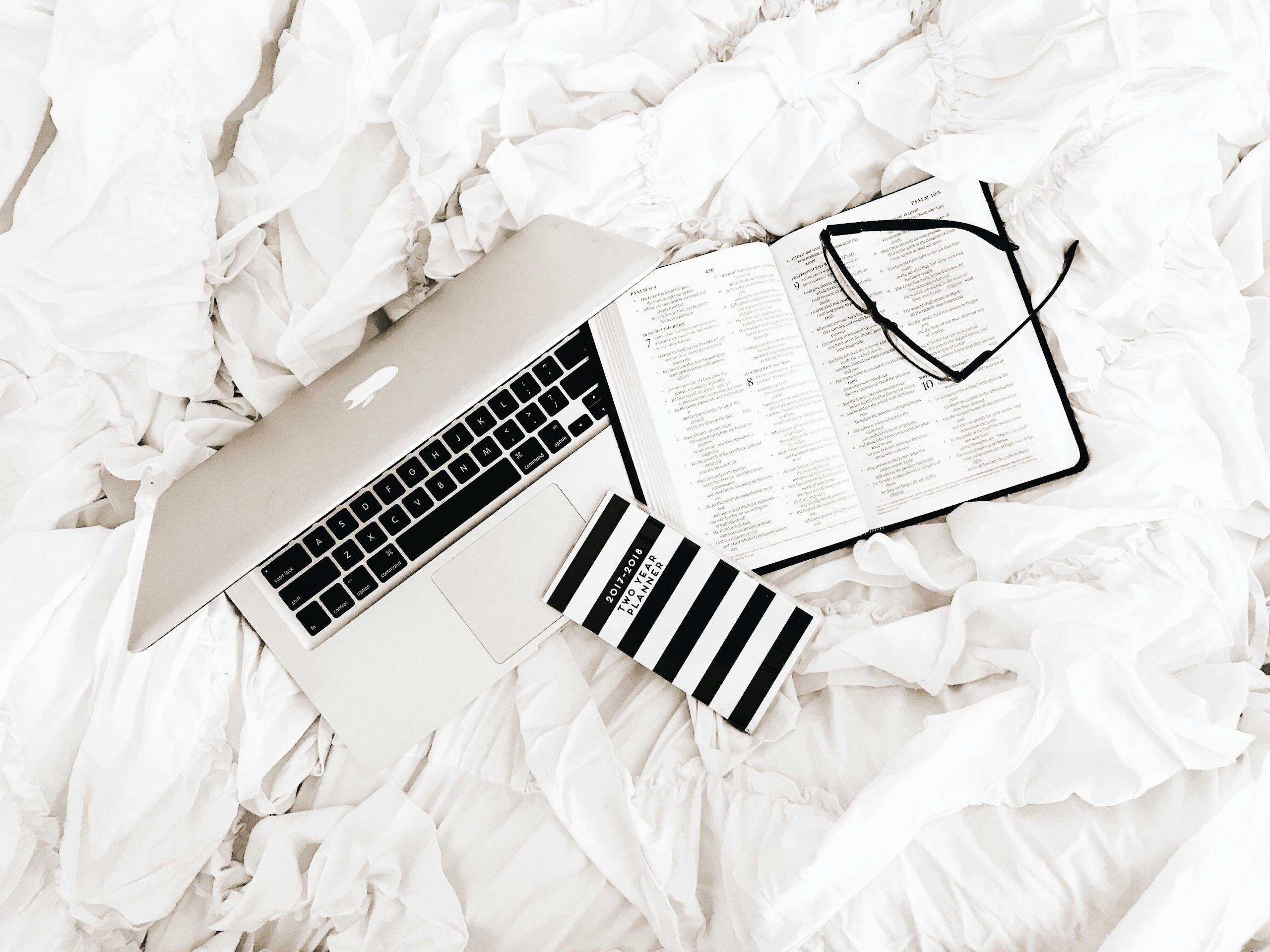 A flat lay of a laptop, open book, and reading glasses on a white bed. Perfect for productivity and relaxation themes.